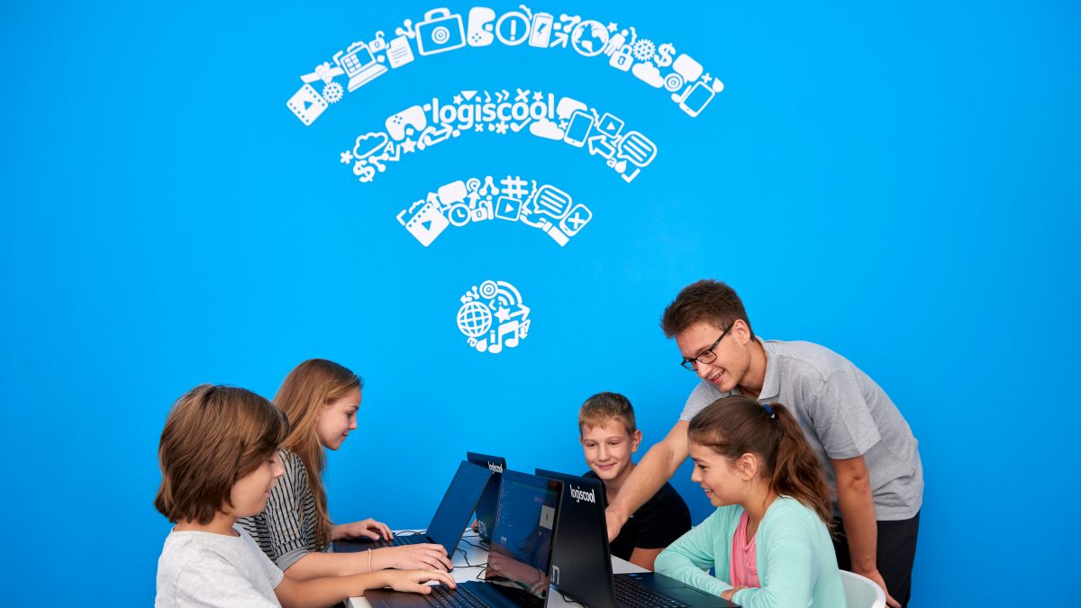 Vier Kinder sitzen vor Laptops und ein junger Mann schaut dabei zu. Der Hintergrund ist leuchtend blau.