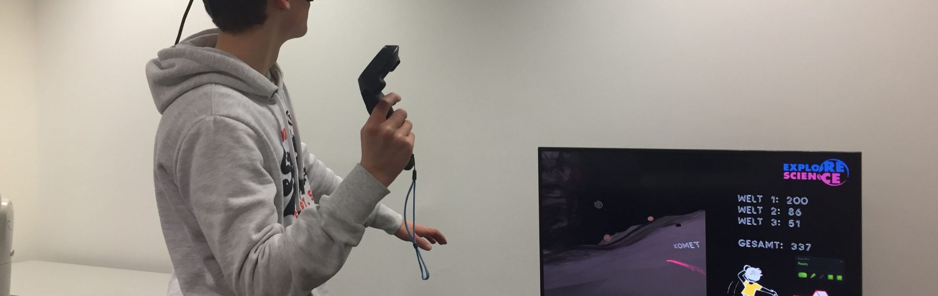 Ein Mann trägt eine Virtual Reality Brille und hat einen Controller in der Hand. Er spielt offensichtlich ein Spiel, wie man auf dem Bildschirm vor ihm sehen kann.