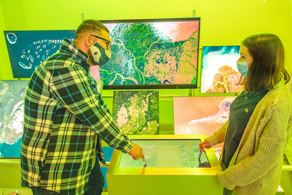 Zwei Personen stehen an einem Touchscreen; im Hintergrund sind Satellitenbilder zu sehen