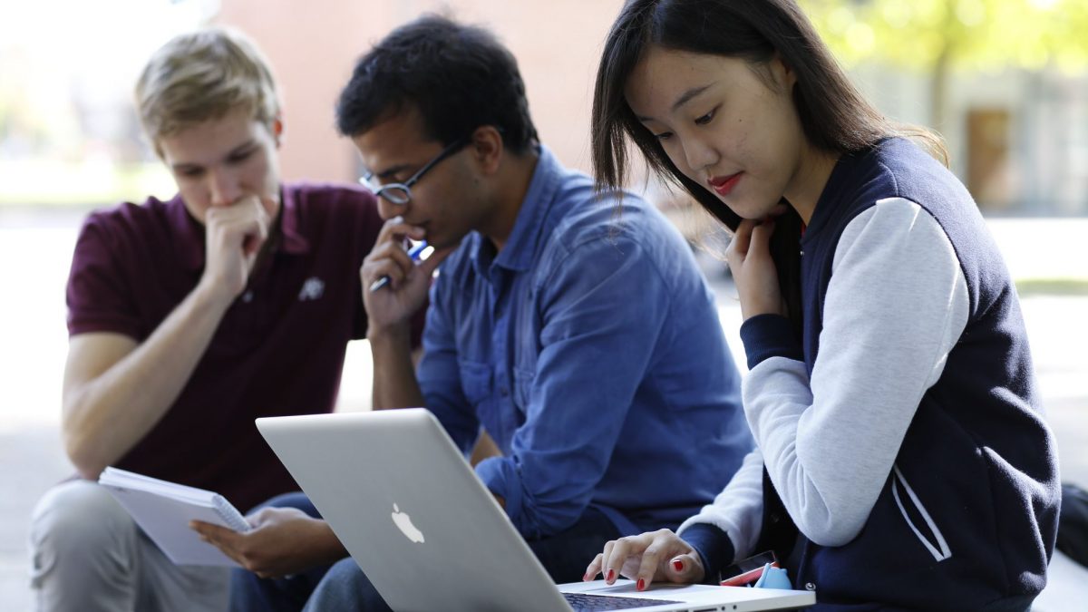Drei junge Menschen sitzen zusammen; Eine schaut in ihren Laptop und zwei auf einen Notizblock