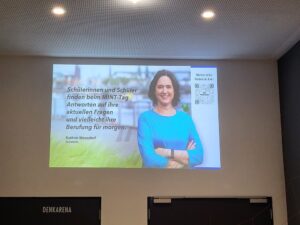 Eine Projektion an der Wand mit einem Zitat von Kathrin Moosdorf, einer Senatorin. Sie spricht über die Bedeutung des MINT-Tages für Schüler und ihre berufliche Orientierung. Ein QR-Code für weitere Informationen ist sichtbar.
